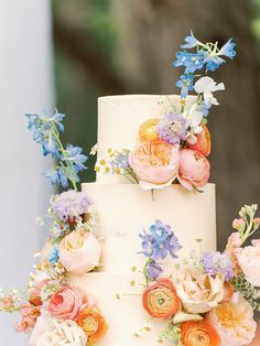 a white wedding cake with flowers on the top and bottom tier is decorated with blue, pink, yellow and orange flowers