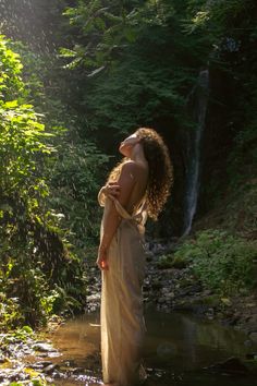 a woman is standing in the water near a waterfall wearing a flowing dress and holding her hair back