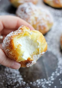 a hand holding a powdered sugar doughnut in front of other pastries on a baking sheet