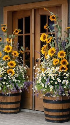 two wooden barrels with sunflowers and other flowers in them on the porch next to a door