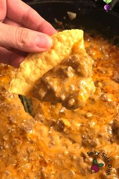 a hand holding a tortilla chip over a skillet filled with chili and cheese