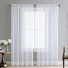 a living room with white curtains and a table in front of the window on top of a rug