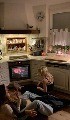 three women sitting on the floor in front of an oven and watching tv at night