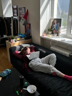 a man laying on top of a black couch in front of a window next to a laptop computer