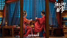 two people dressed in traditional chinese garb sitting at a table with blue drapes