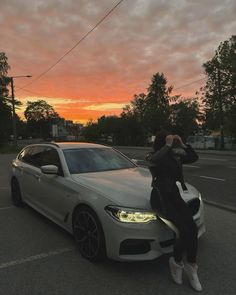 a woman taking a photo next to a white car at sunset with the sun setting in the background