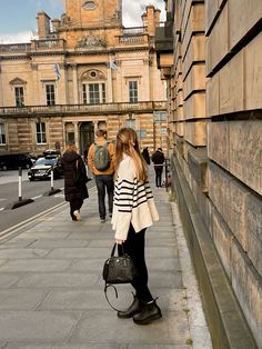 a woman standing on the side of a street holding a black purse and looking down