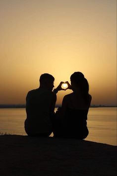 a man and woman sitting on the beach at sunset making a heart shape with their hands