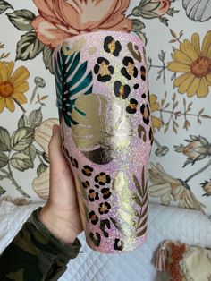 a hand holding a pink and gold leopard print tumbler cup in front of a floral wallpaper