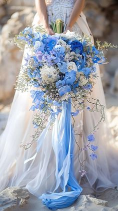 a bridal holding a blue and white bouquet on the beach with rocks in the background