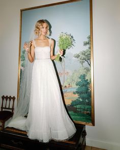 a woman standing in front of a painting wearing a wedding dress and holding a bouquet of flowers