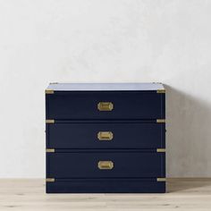 a blue chest with brass handles on a wooden floor next to a white wall and lamp