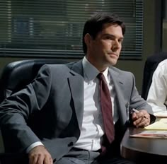 two men in suits sitting at a table with papers on it and one man wearing a tie