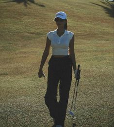 a woman walking across a grass covered field holding a golf club and wearing a white shirt