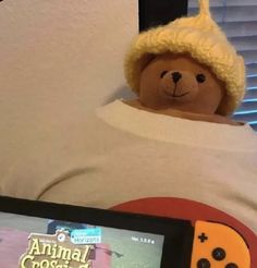 a teddy bear wearing a yellow hat and holding a nintendo wii game controller in its lap