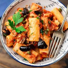 a bowl filled with pasta and olives on top of a wooden table next to a fork