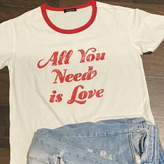 All You Need Is Love Graphic Tee White Casual Top For Valentine's Day, Casual Red Tops For Valentine's Day, Casual Valentine's Day Top With Text Print, Casual White Top For Valentine's Day, White Graphic Tee For Valentine's Day, Red Graphic Tee For Valentine's Day, White Casual T-shirt For Valentine's Day, Casual Valentine's Day Graphic T-shirt, Valentine's Day Graphic Tee T-shirt