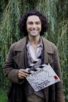 a man holding a movie clapper in front of some trees and bushes, smiling at the camera