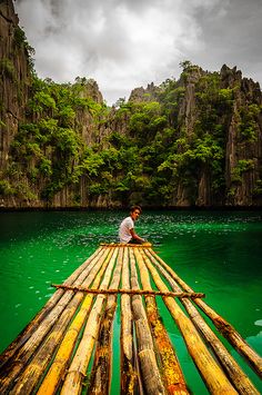 a man sitting on top of a bamboo raft in the middle of a lake surrounded by mountains