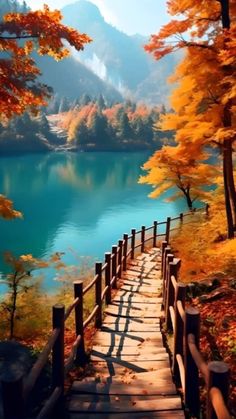 a wooden walkway leading to a lake surrounded by autumn leaves and trees with mountains in the background