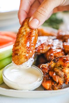 a person dipping sauce on chicken wings with celery and cucumbers in the background