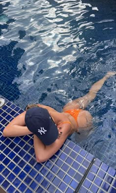 a woman in an orange swimsuit and hat sitting on the edge of a swimming pool