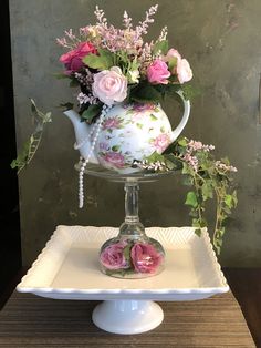 a white tea pot with pink flowers in it sitting on a plate next to a flower arrangement