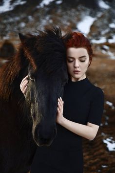 a woman with red hair is hugging a black horse's head in the snow