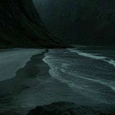 an empty beach with mountains in the background and dark clouds over water on either side