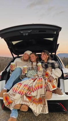 three women sitting in the back of a car with their blankets on and holding coffee cups