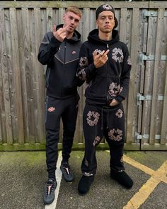 two young men standing next to each other in front of a wooden fence wearing matching tracksuits