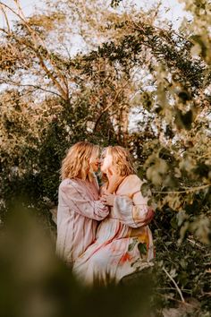 two women hugging each other in front of some trees and bushes, one is wearing a pink dress