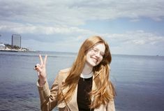 a woman with long red hair and glasses making the peace sign by the water's edge