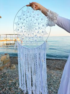 a woman holding up a white dream catcher on the beach with water in the background