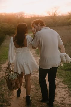 a man and woman walking down a dirt road holding hands while the sun is setting