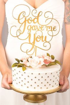 a woman holding a white cake with gold lettering on top and flowers in the bottom