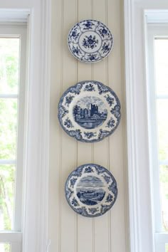 three blue and white plates hanging on the wall next to each other in front of two windows