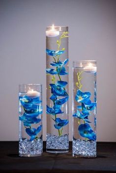 three tall glass vases with blue flowers and lit candles in them on a table