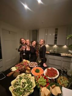 three women standing in front of a table full of food and drinks with wine glasses