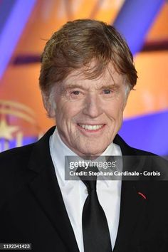 an older man in a black suit and tie smiles at the camera while posing for a photo