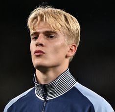 a young man with blonde hair wearing a blue and white jacket looking off into the distance