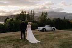 a bride and groom standing in front of an old car on top of a hill