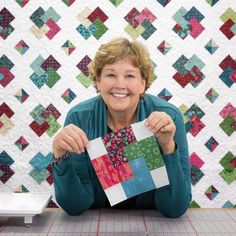 a woman sitting at a table holding up a piece of quilting material with the background made from squares