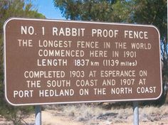 a sign that is in front of some bushes and trees with writing on it saying, no 1 rabbit proof fence the largest fence in the world