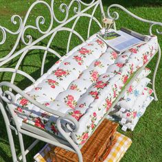 a white iron bench with flowers on it in the grass next to a picnic basket