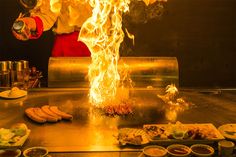 a chef is cooking food with flames in the air and on fire behind him are other dishes