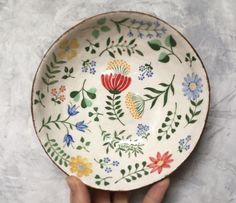 a hand holding a bowl with flowers and leaves painted on the side, in front of a white background