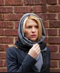 a woman wearing a gray scarf and black jacket standing in front of a brick wall