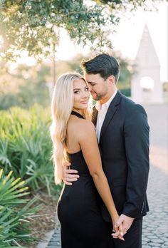 a man and woman standing next to each other in front of a tree with their arms around each other