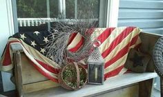 an american flag is draped over a wooden bench on the front porch, with a lantern and potted plant